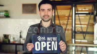 开朗英俊的男人小咖啡馆老板拿着`我们在咖啡店里打开`标志。 开幕